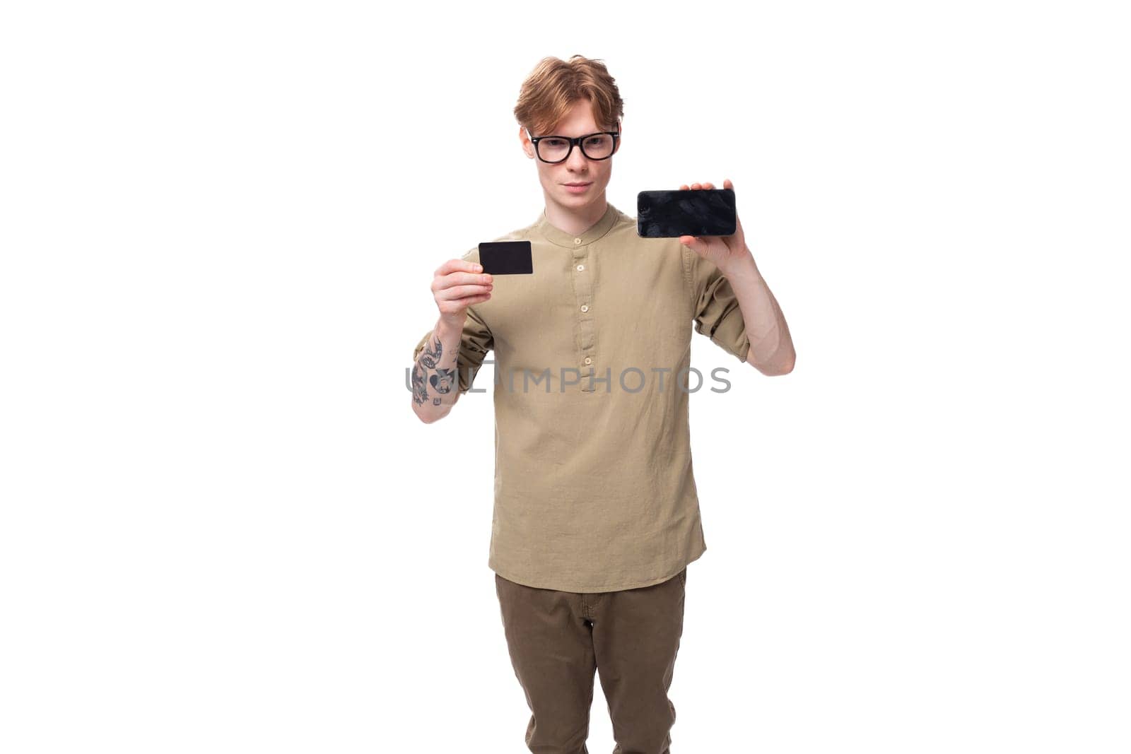 handsome young caucasian male promoter with red hair and a tattoo on his arm dressed in a khaki shirt holds a credit card and a phone.