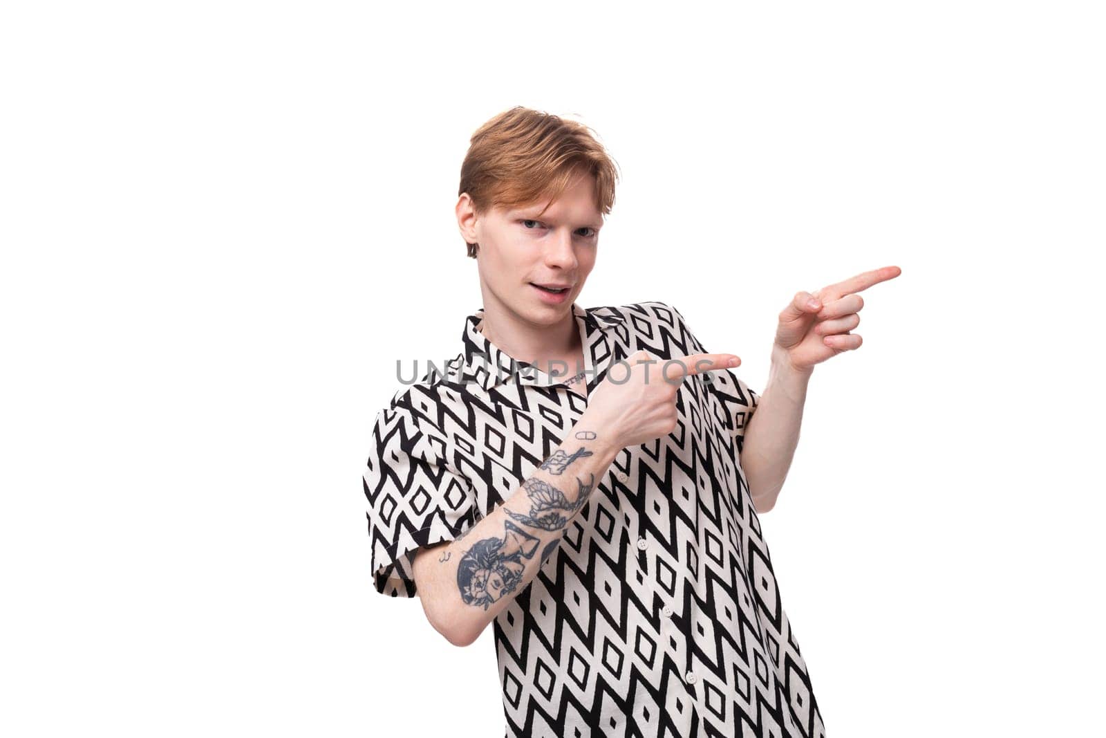 a young handsome guy with red hair and a tattoo on his arms, dressed in a short-sleeved shirt, points with his hand to the space for advertising. business concept.