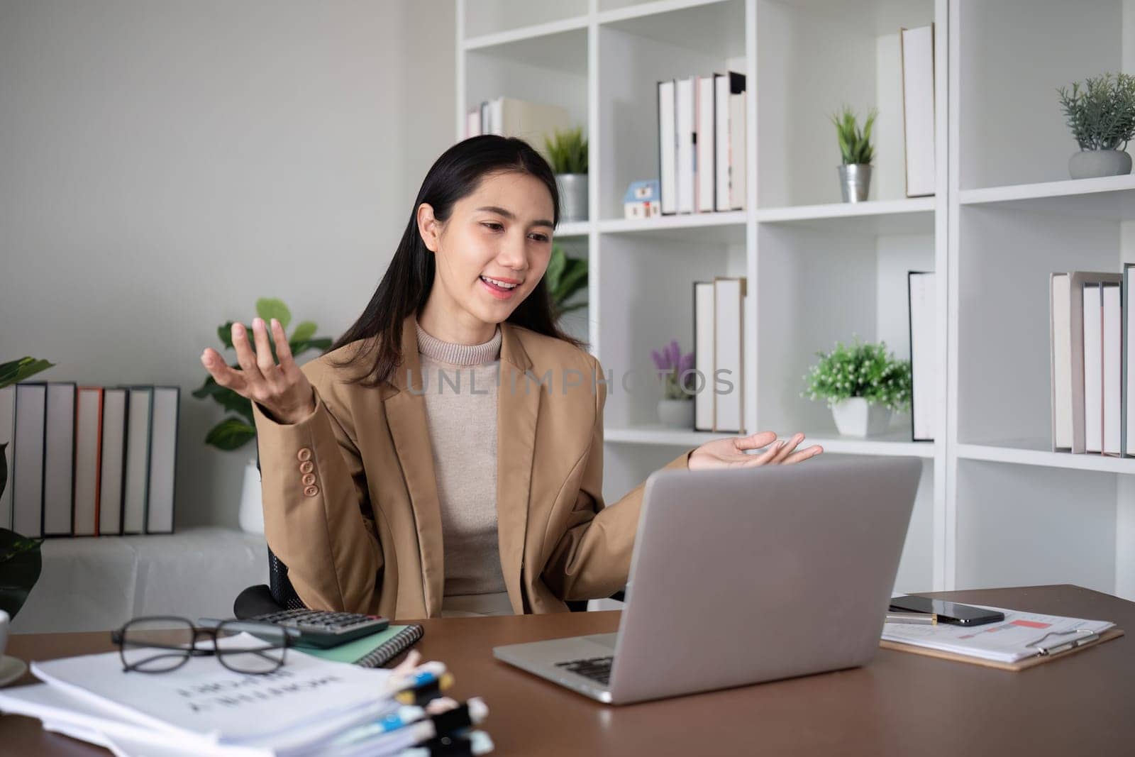 Young Asian business woman sits and works using a laptop in a modern office decorated with shady green plants..