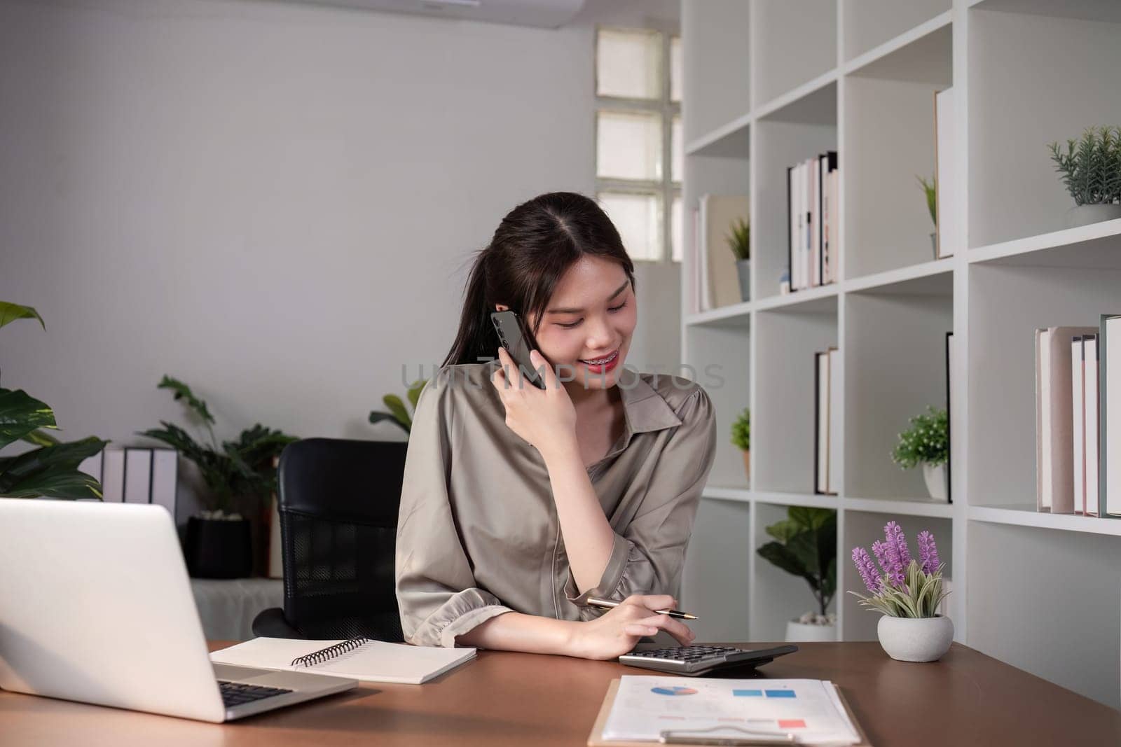 Young Asian business woman sits on the phone in an online business meeting using a laptop in a modern home office decorated with shady green plants..