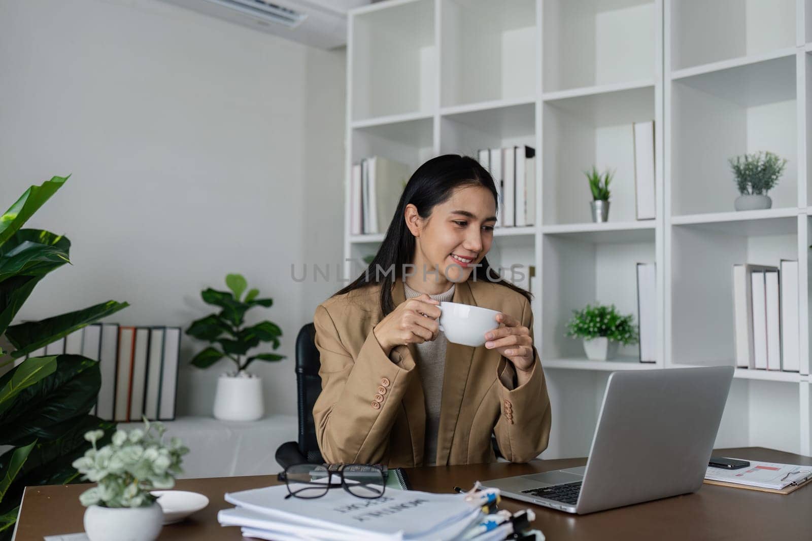 smiling businesswoman drinking coffee and working on laptop in green modern office. Nice Environment Office. Eco-Friendly Concept by itchaznong