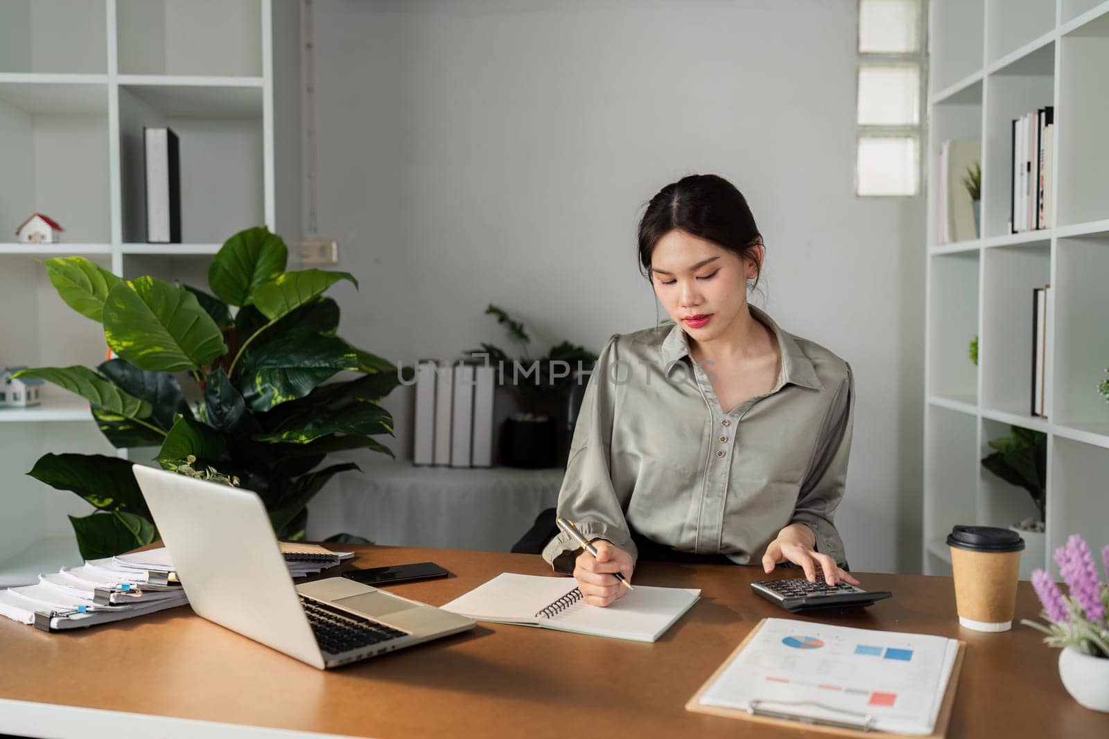 A woman is sitting at a desk with a calculator and a pen. She is writing in a notebook
