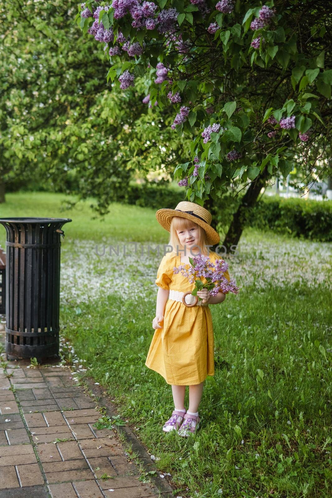 A little girl in a yellow dress and straw hat wearing a bouquet of lilacs. A walk in a spring park, blossoming lilacs