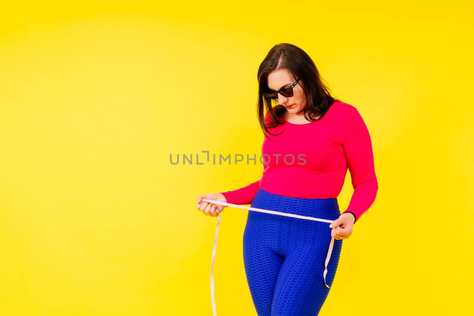 Pretty female with excess weight in sporty top measuring waist over white yellow background by Zelenin