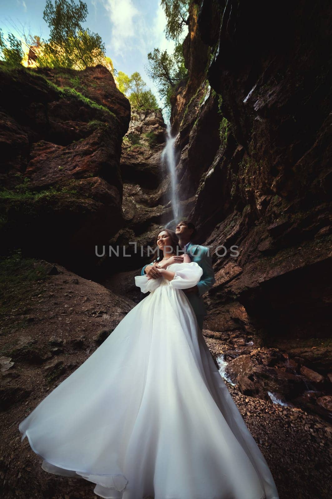 Couple in love against the backdrop of a waterfall. Honeymoon trip. Happy couple in the mountains. Vertical photo of newlyweds hugging against the backdrop of a waterfall in the gorge by yanik88