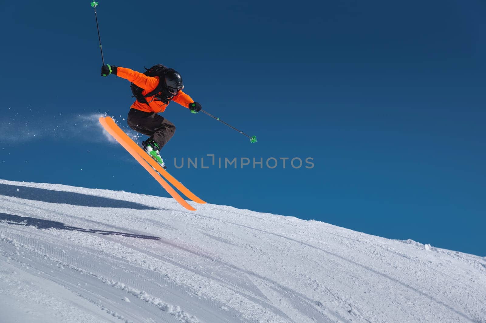 A skier in an orange jacket and a black helmet makes a jump on a sunny day against a clear sky. Copy space for your information by yanik88