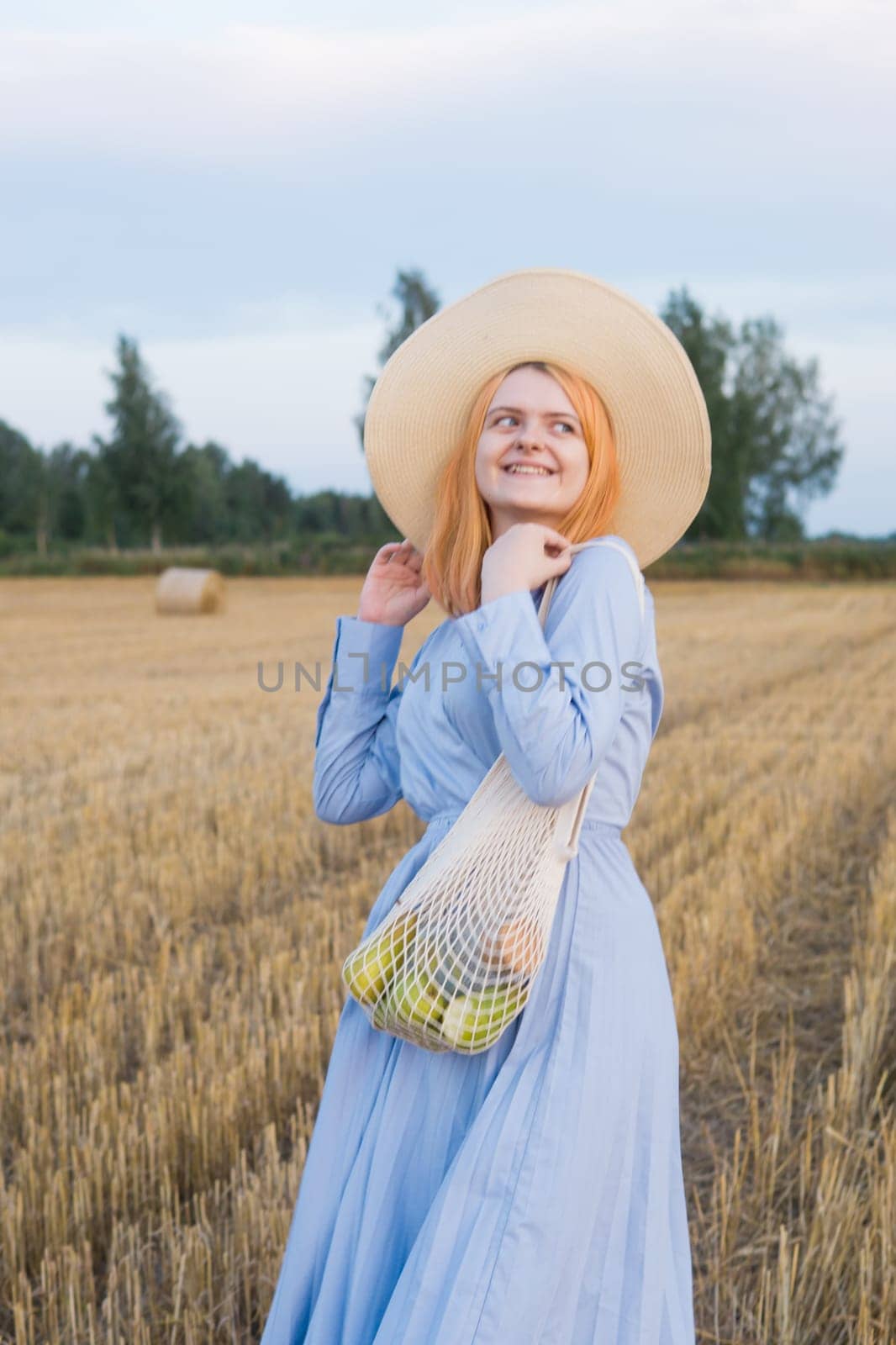 A red-haired woman in a hat and a blue dress walks in a field with haystacks