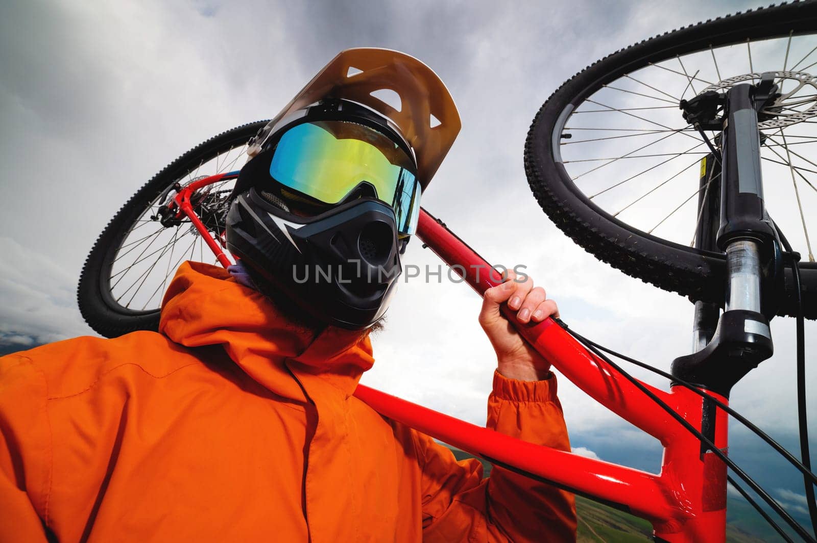Wide angle vertical frame top angle. Portrait of a bearded mountain biker with his bicycle against a background of mountains and clouds in the summer surrounded by green grass.