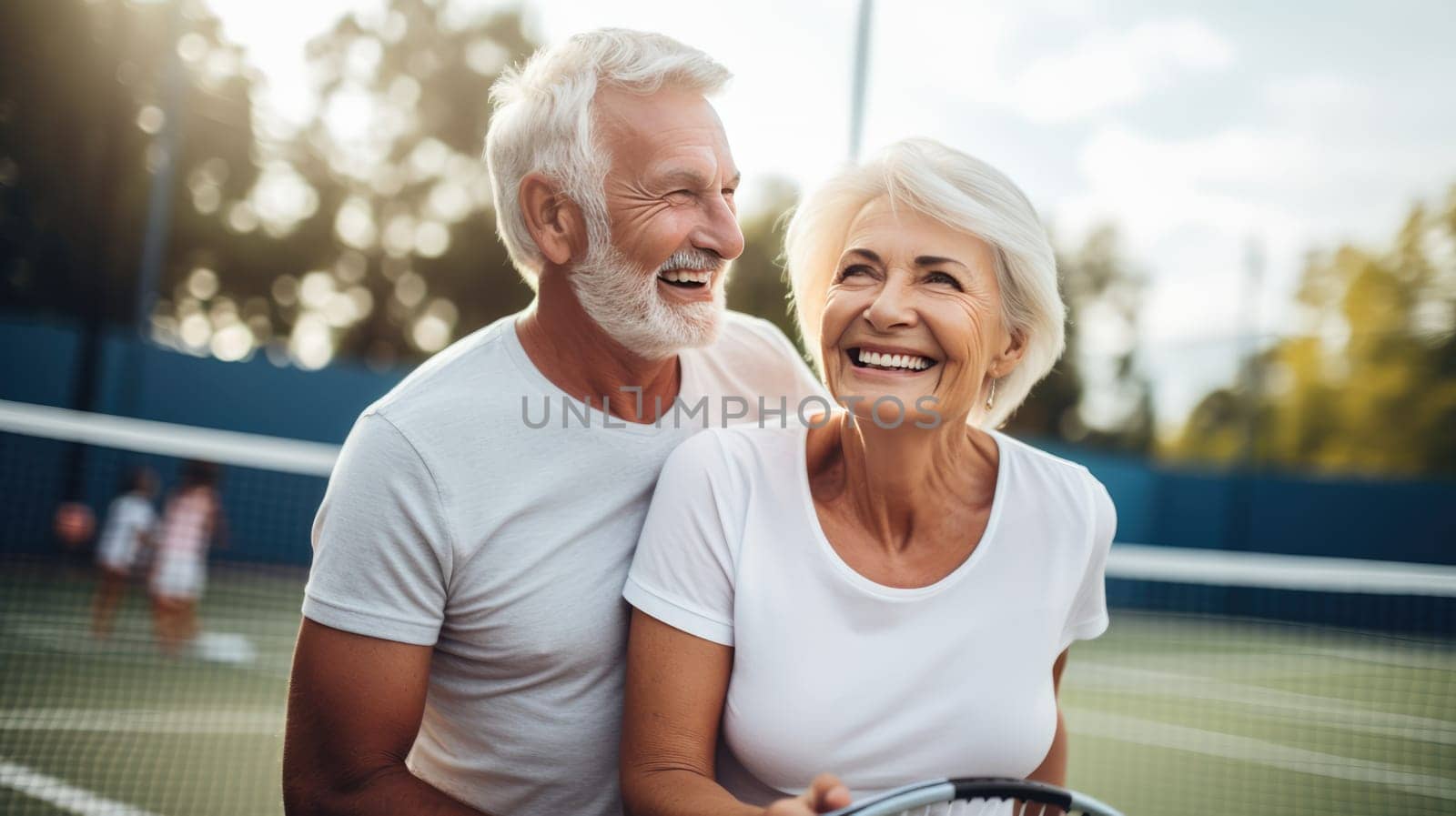 Horizontal shot of a happy elderly couple standing closely together on a tennis court and smiling at each other. They both have sporty outfits on and the man is wearing a tennis cap.