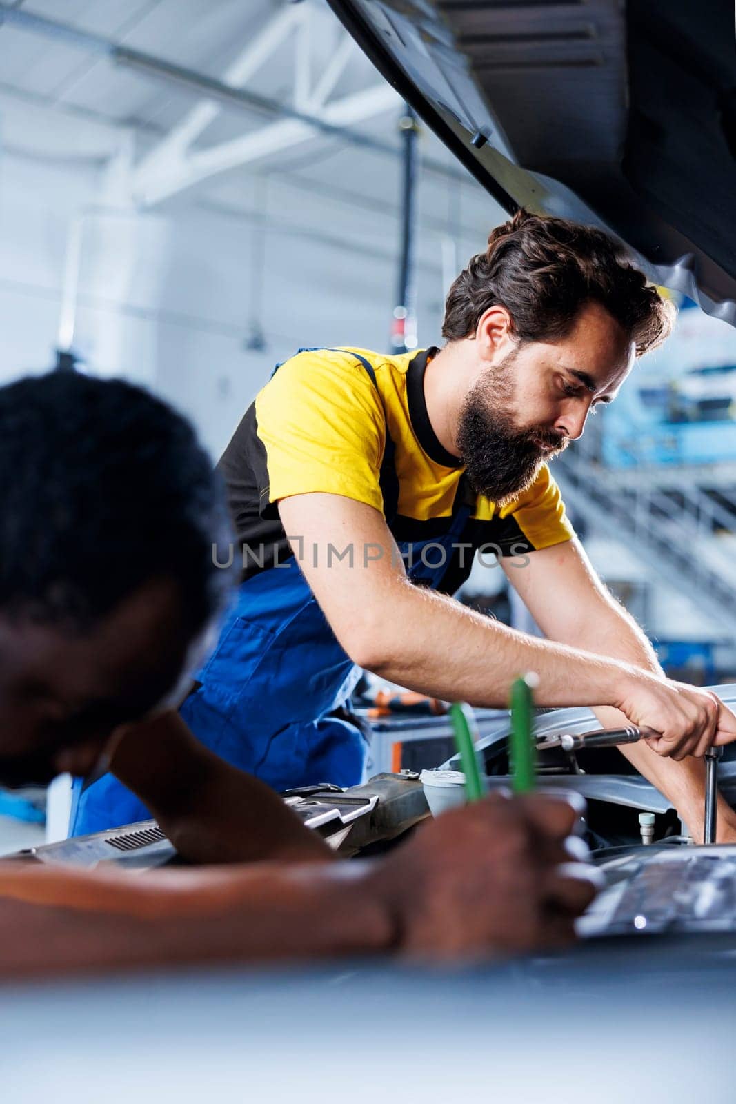 Repairman and coworker collaborating on servicing broken vehicle, checking for faulty motor. Licensed mechanics in auto repair shop working together on fixing car, discussing best options