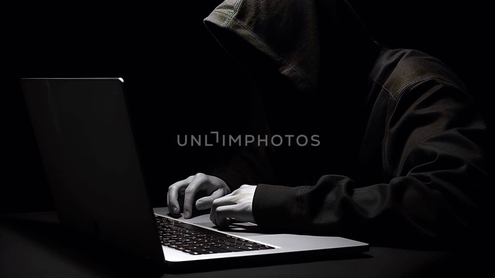 An obscured figure in a hoodie types on a laptop in the dark, evoking themes of cybercrime and online security threats by chrisroll