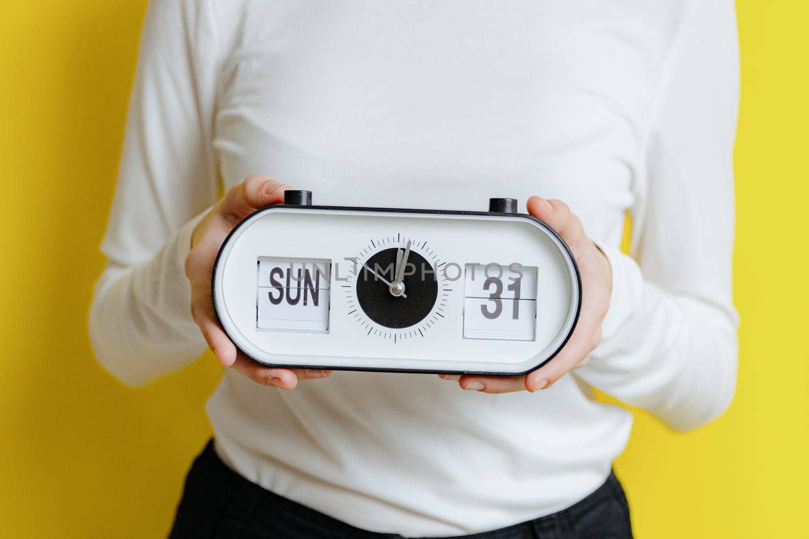 The hands of one young Caucasian unrecognizable girl holds a clock with time, date and day: Sunday, lunch, 31, standing on a yellow background on a spring day in the room, side view close-up.