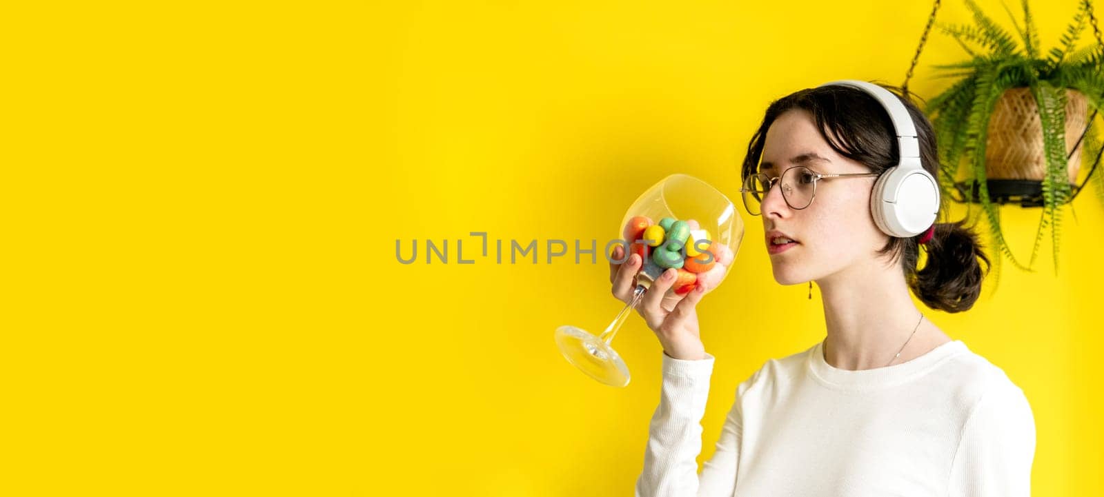 One young Caucasian beautiful teenage girl in glasses and headphones holds a wine glass with Easter marble decorative eggs standing and looking to the side to the right on a yellow background with a hanging palm flower on a spring day in a room with copy space on the left, side view close-up.