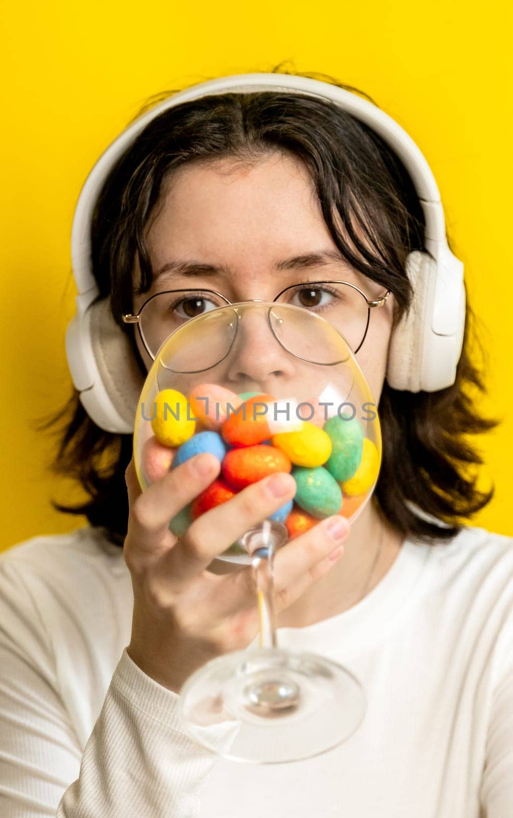 Portrait of one young Caucasian beautiful teenage girl in glasses and headphones drinks from a wine glass with Easter marble decorative eggs on a yellow background on a spring day in the room and looks at the camera, side view close-up with selective focus.