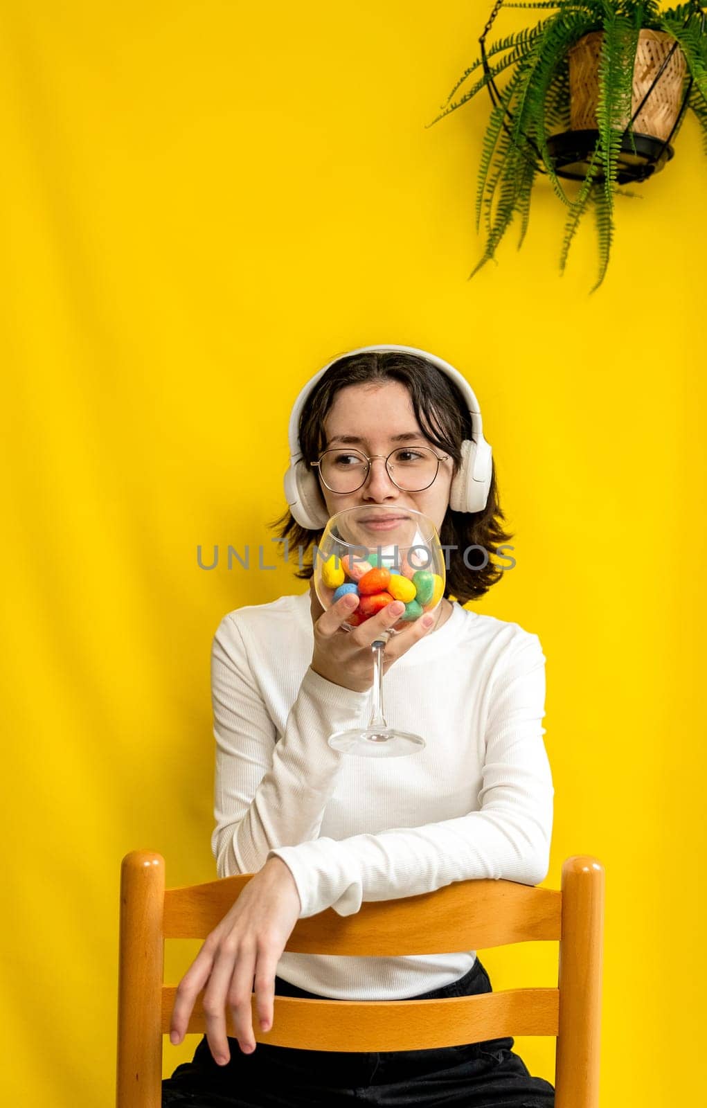 Portrait of one young Caucasian beautiful happy teenage girl in glasses and headphones holds a wine glass with Easter marble decorative eggs at her mouth, sitting on a chair on a yellow background with a hanging palm flower on a spring day in the room and looks to the side, side view close-up.