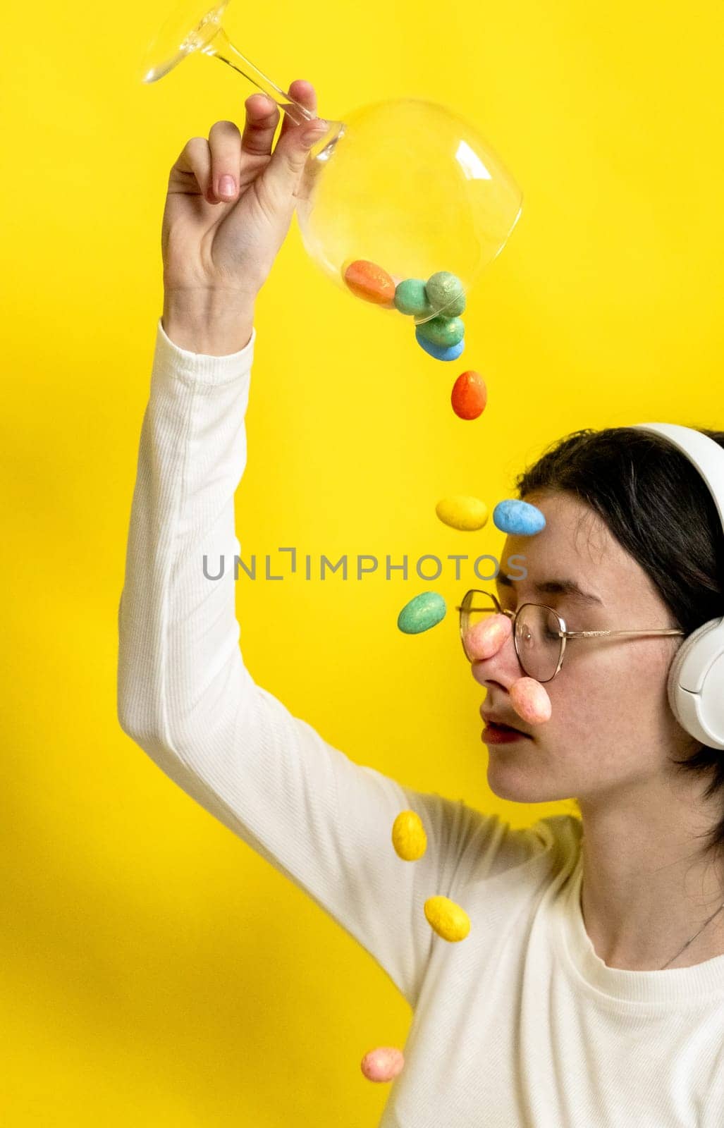 Portrait of one young Caucasian beautiful happy teenage girl with closed eyes in glasses and headphones pours out of a wine glass with Easter marble decorative eggs onto her face, sitting sideways on a yellow background on a spring day in the room, side view close-up.
