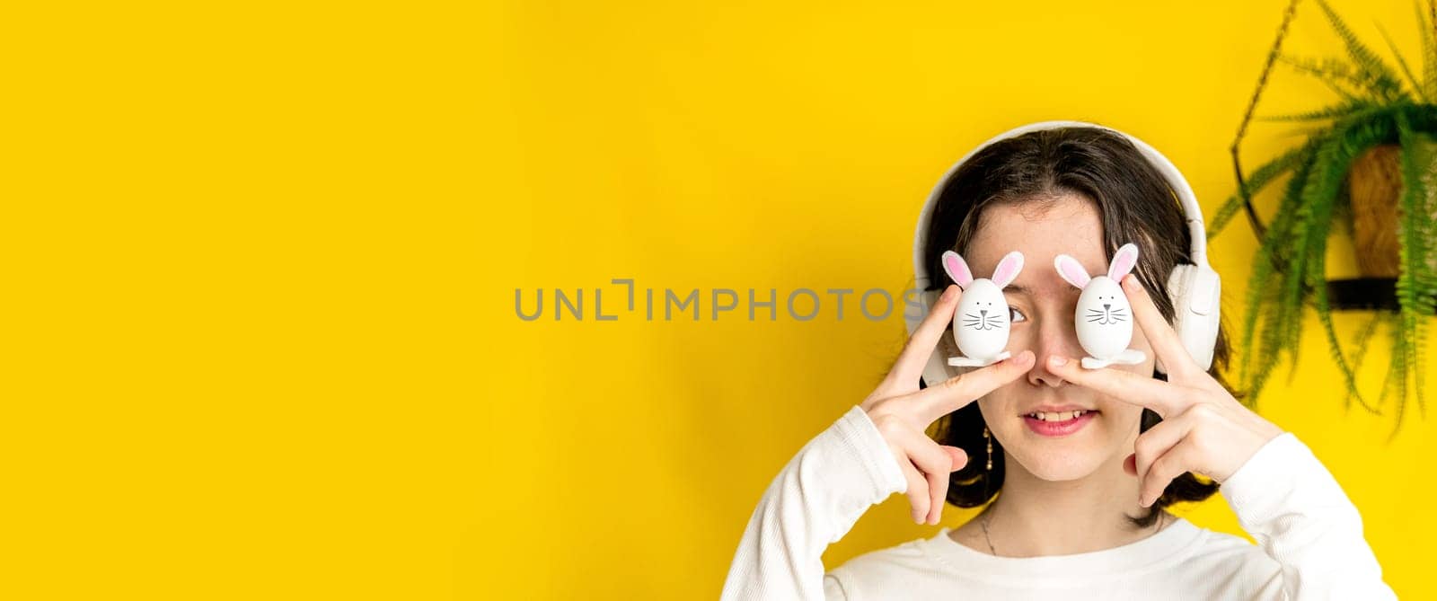 Portrait of one young Caucasian beautiful happy teenage girl holding with her fingers two Easter decorative bunny at eye level, standing on the right on a yellow background with a hanging palm flower on a spring day in a room with copy space on the left, side view close-up.