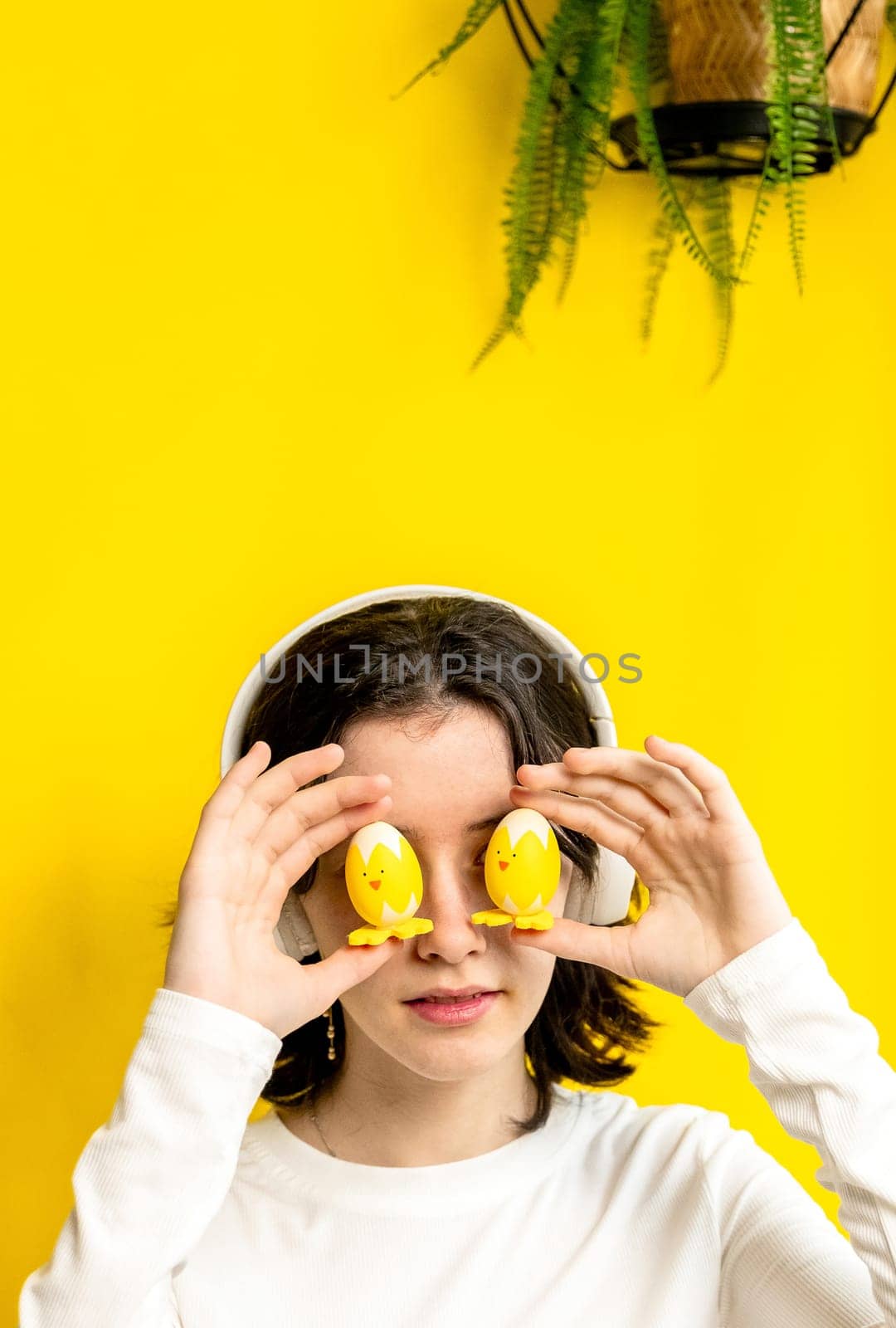 Portrait of one young Caucasian beautiful happy teenage girl holding with her fingers two Easter decorative chickens at eye level, standing on a yellow background with a hanging palm flower on a spring day in a room with small copy space, side view close-up.