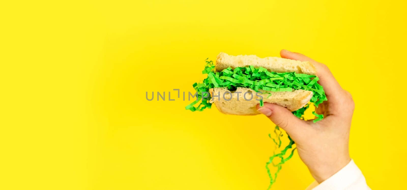 Portrait of one hand of a young Caucasian teenage girl holding a bread sandwich on the right with Easter decorative green hay on a yellow background with with copy space on the left on a spring day in the room, side view close-up with depth of field.
