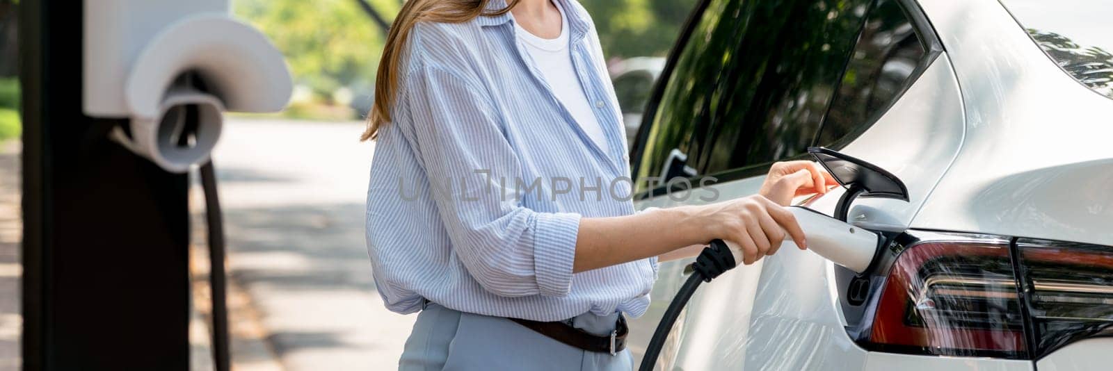 Young woman recharging battery for electric car during road trip travel EV car in natural forest or national park. Eco friendly travel during vacation and holiday. Panorama banner Exalt