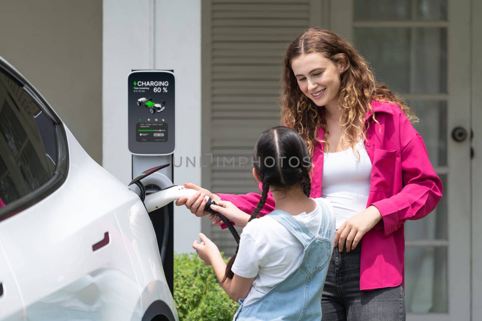 Happy little young girl learn about eco-friendly and energy sustainability as she help her mother recharge electric vehicle from home EV charging station. EV car and modern family concept. Synchronos