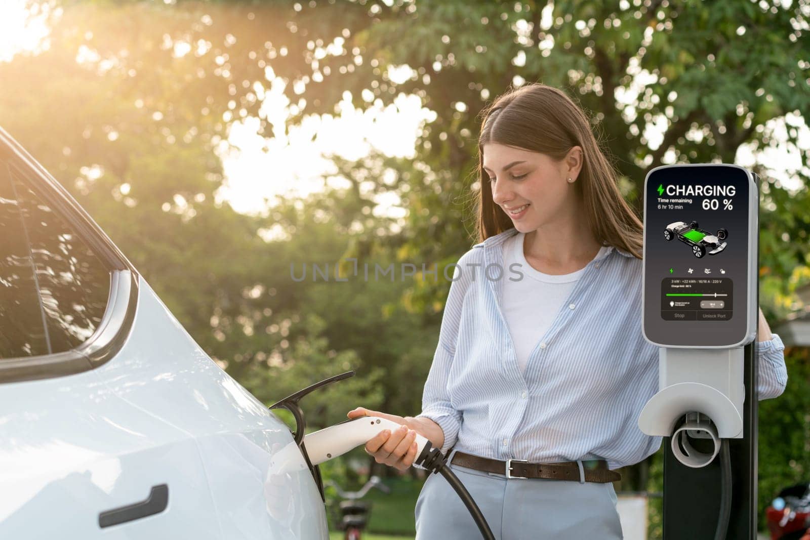 Young woman recharging battery for electric car during road trip travel EV car in natural forest or national park with sunrise lighting. Eco friendly travel during vacation and holiday. Exalt