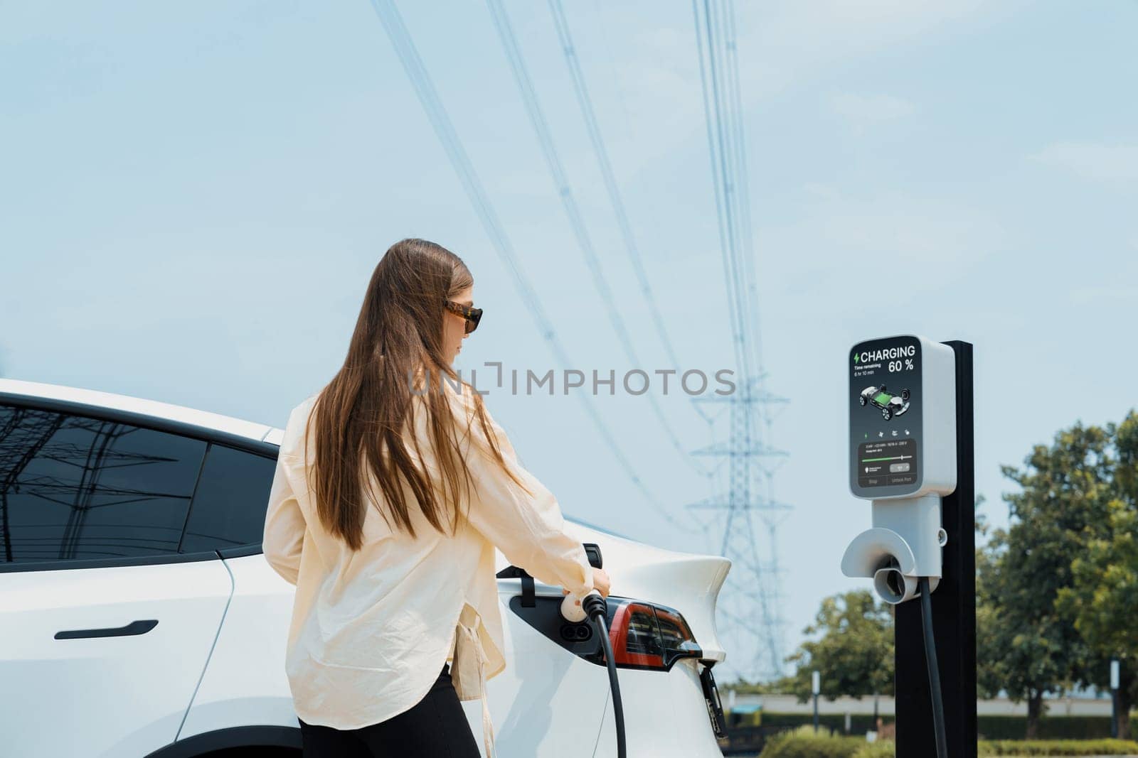 Young woman recharge EV car battery at charging station connected to power grid tower electrical industrial facility as electrical industry for eco friendly vehicle utilization. Expedient
