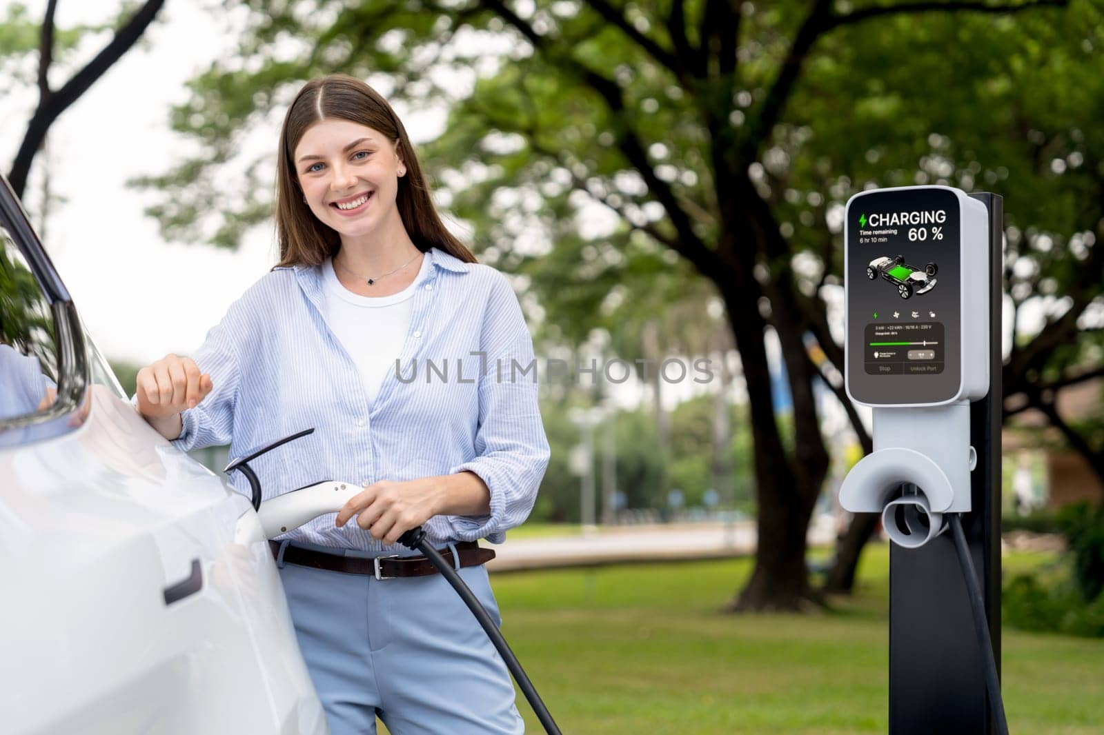 Young woman recharge EV electric vehicle's battery from EV charging station in outdoor green city park scenic. Eco friendly urban transport and commute with eco friendly EV car travel. Exalt