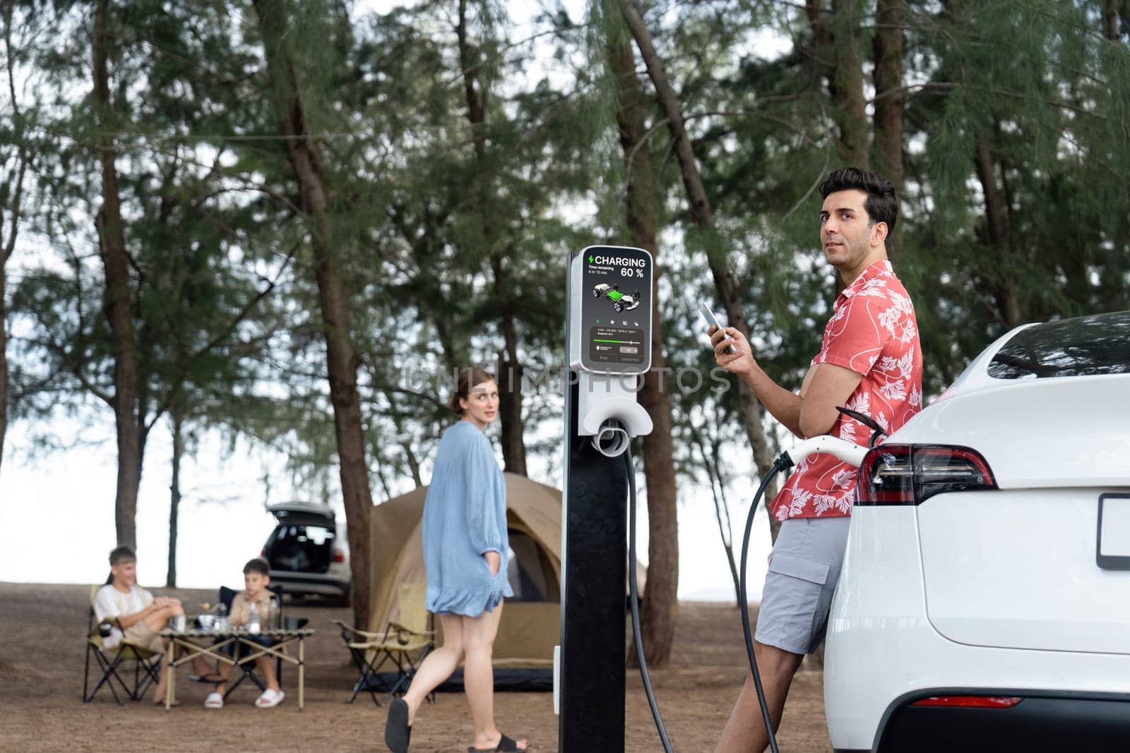 Outdoor adventure and family vacation camping in nature travel by eco friendly car for sustainable future. Lovely sit on trunk, charging EV car with EV charging station in campsite. Perpetual