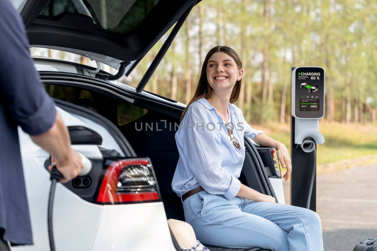 Lovely young couple recharging battery for electric car while relaxing and sitting on the trunk during road trip travel EV car in autumnal forest. Eco friendly travel on vacation during autumn. Exalt