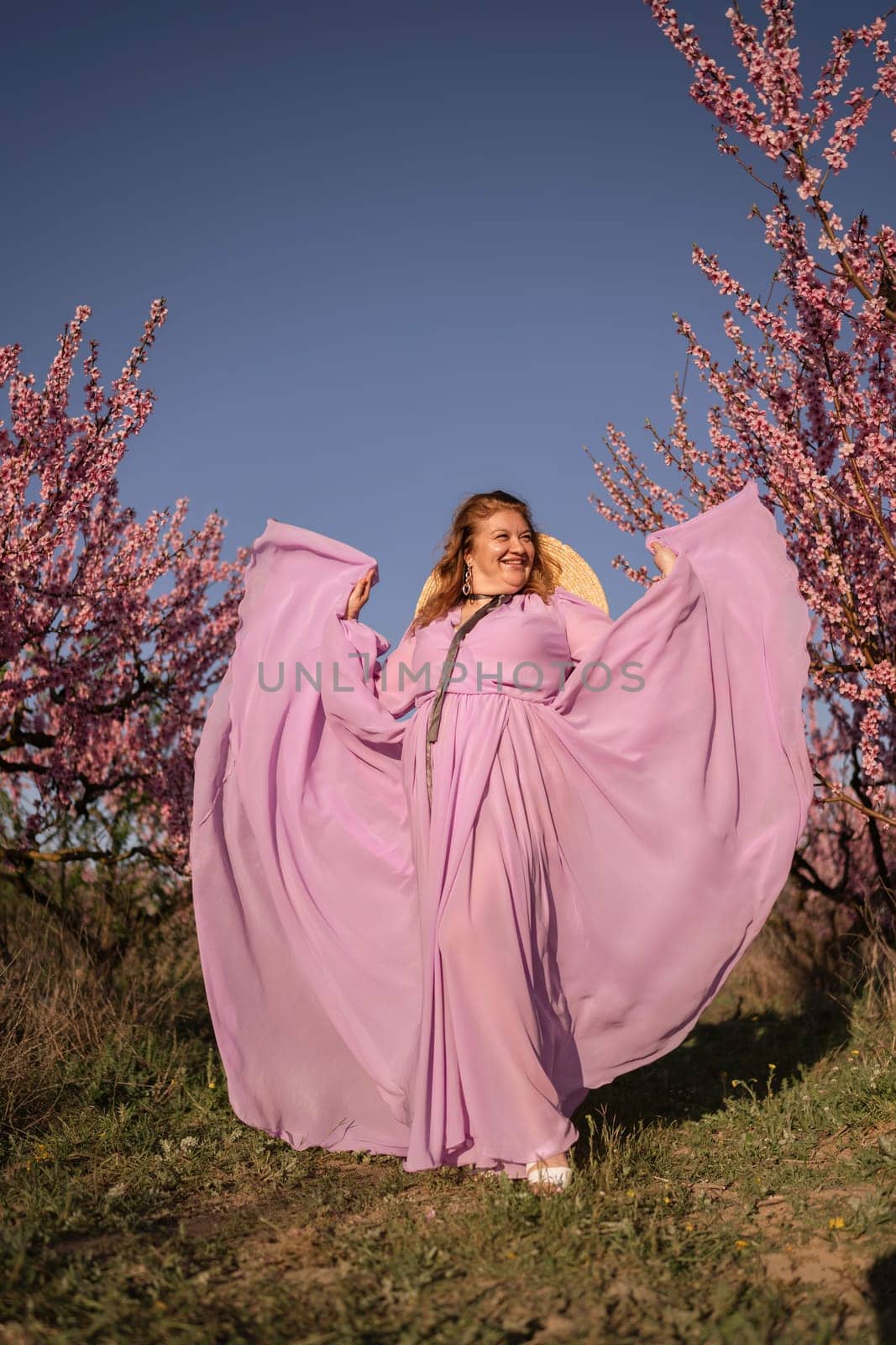 Woman blooming peach orchard. Against the backdrop of a picturesque peach orchard, a woman in a long pink dress and hat enjoys a peaceful walk in the park, surrounded by the beauty of nature