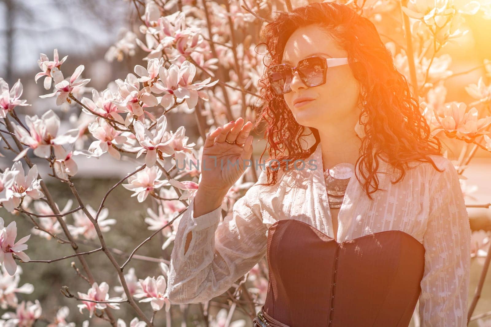 Magnolia park woman. Stylish woman in a hat stands near the magnolia bush in the park. Dressed in white corset pants and posing for the camera. by Matiunina