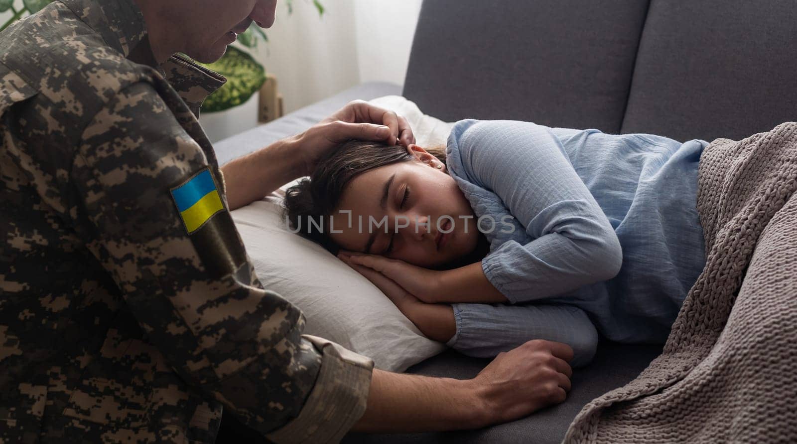 Soldier in Ukrainian military uniform kissing his daughter while she sleeping on sofa at home. Family reunion
