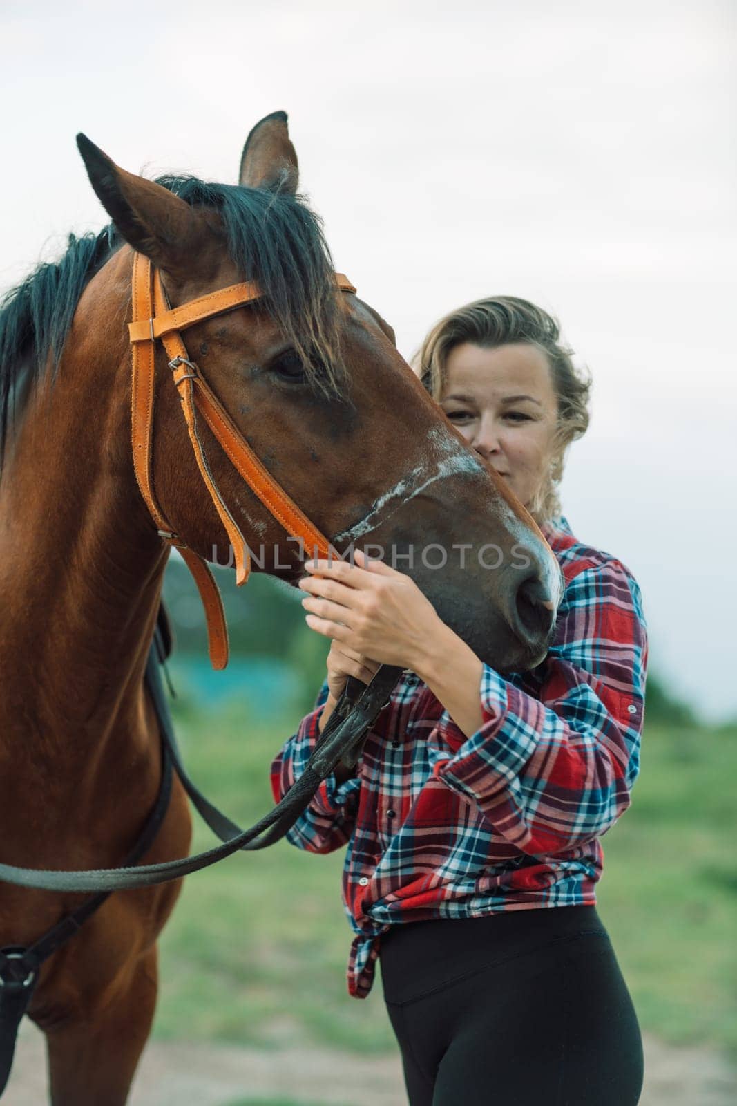 Happy blonde with horse in forest. Woman and a horse walking through the field during the day. Dressed in a plaid shirt and black leggings