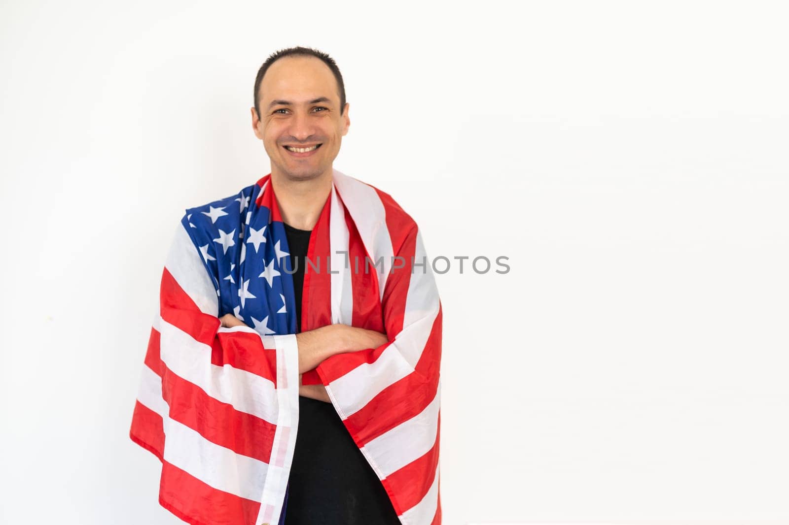 Happy relaxed satisfied handsome middle aged man standing wrapped in USA flag, celebrating national holiday, wearing urban style hoodie. Indoor studio shot isolated on white background. by Andelov13