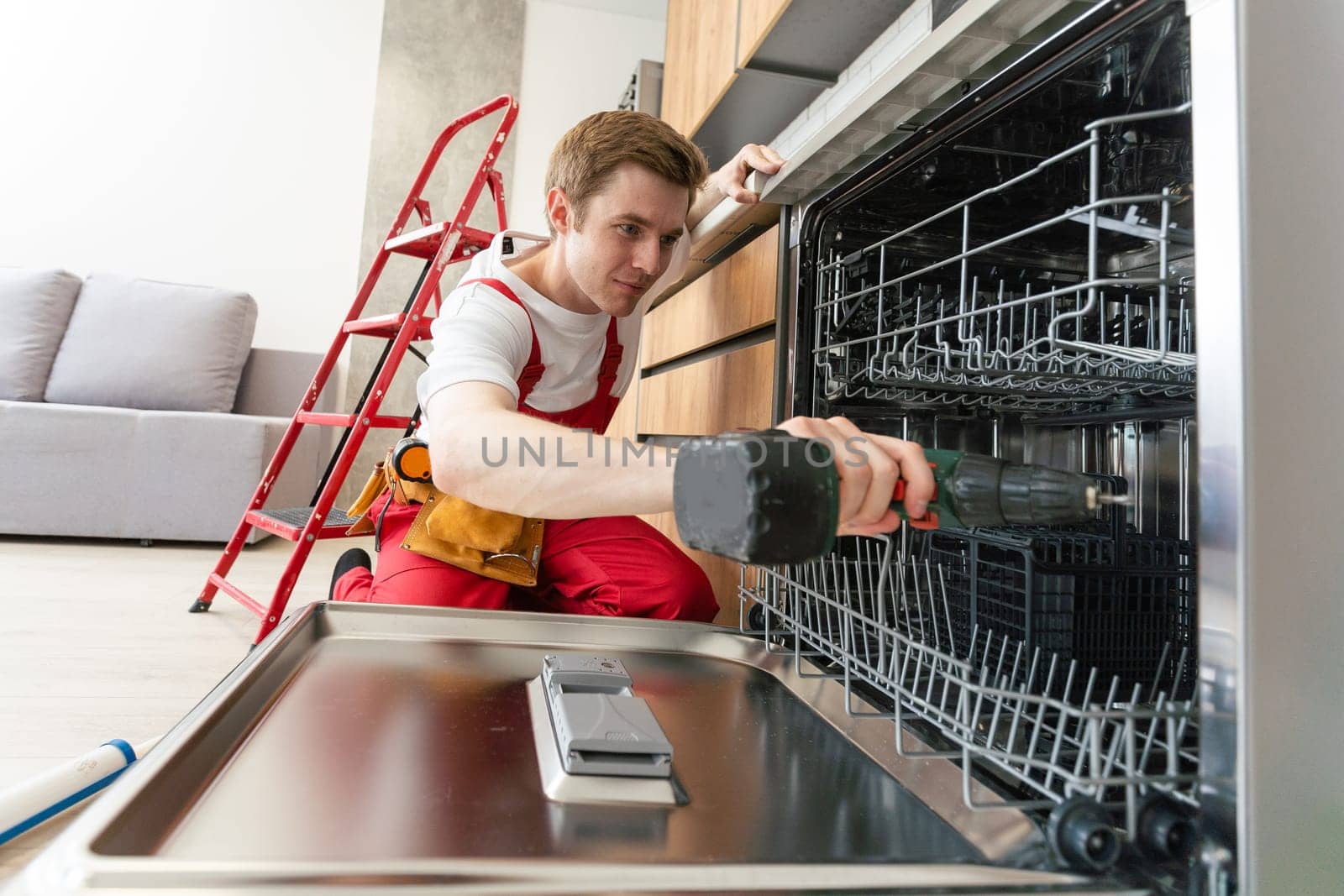 Technician or worker in uniform installs dishwasher into the kitchen furniture. Repairman wear worker suit repairing maintenance of dishwasher. Master in protective gloves fix dishwasher.