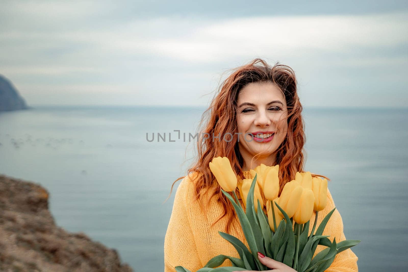 Portrait of a happy woman with hair flying in the wind against the backdrop of mountains and sea. Holding a bouquet of yellow tulips in her hands, wearing a yellow sweater by Matiunina