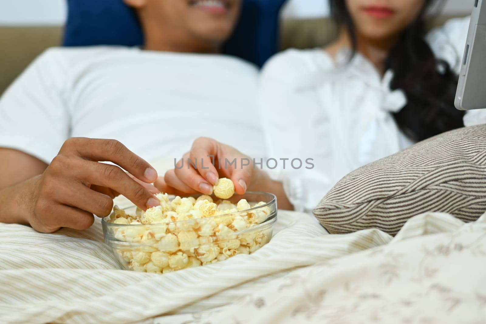 Cropped shot of young couple eating popcorn and watching movie on bed at home.
