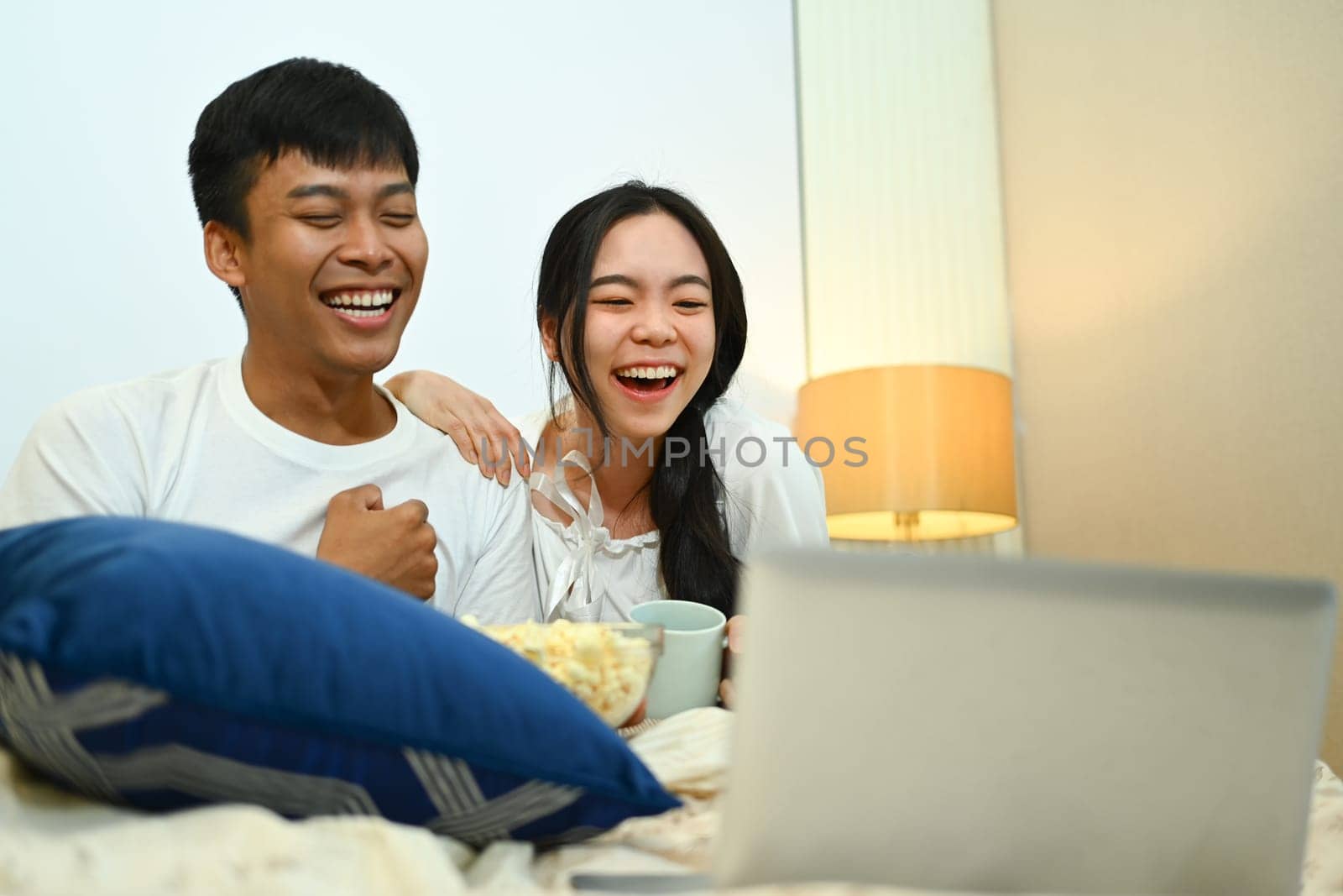 Happy young couple watching movie on laptop and eating popcorn in their bedroom.