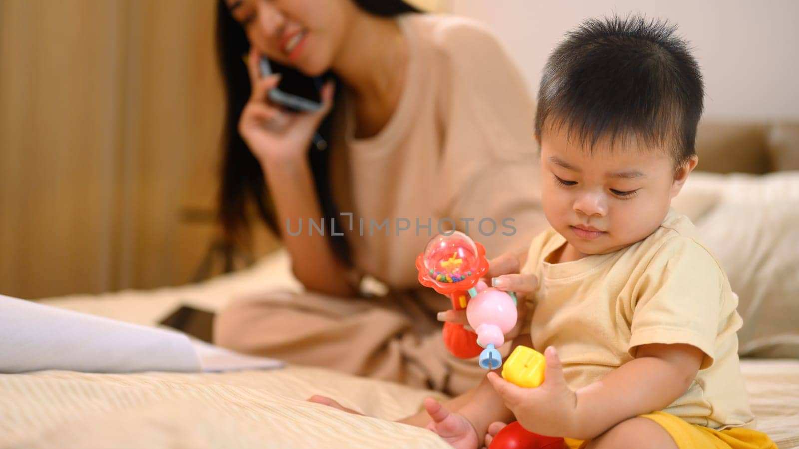 Busy young mother talking on cellphone and little baby boy playing with toy near her.