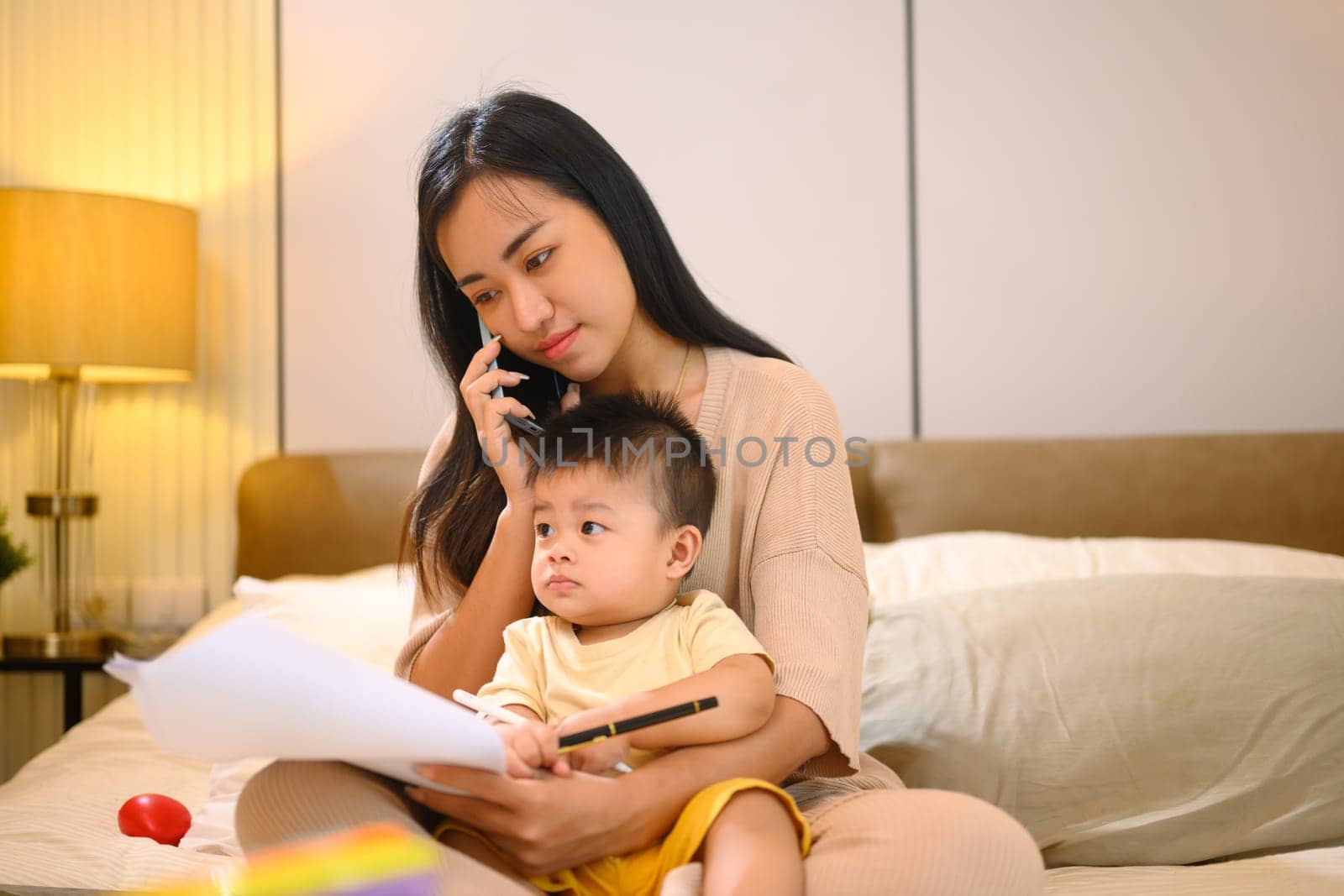 Busy working mom talking on mobile phone and holding her baby son sitting on bed.
