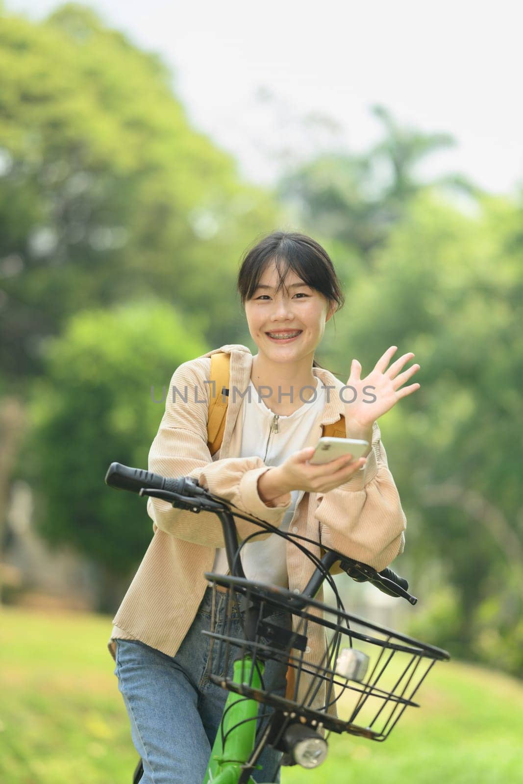 Portrait of cheerful young female student sitting on bicycle and using mobile phone by prathanchorruangsak