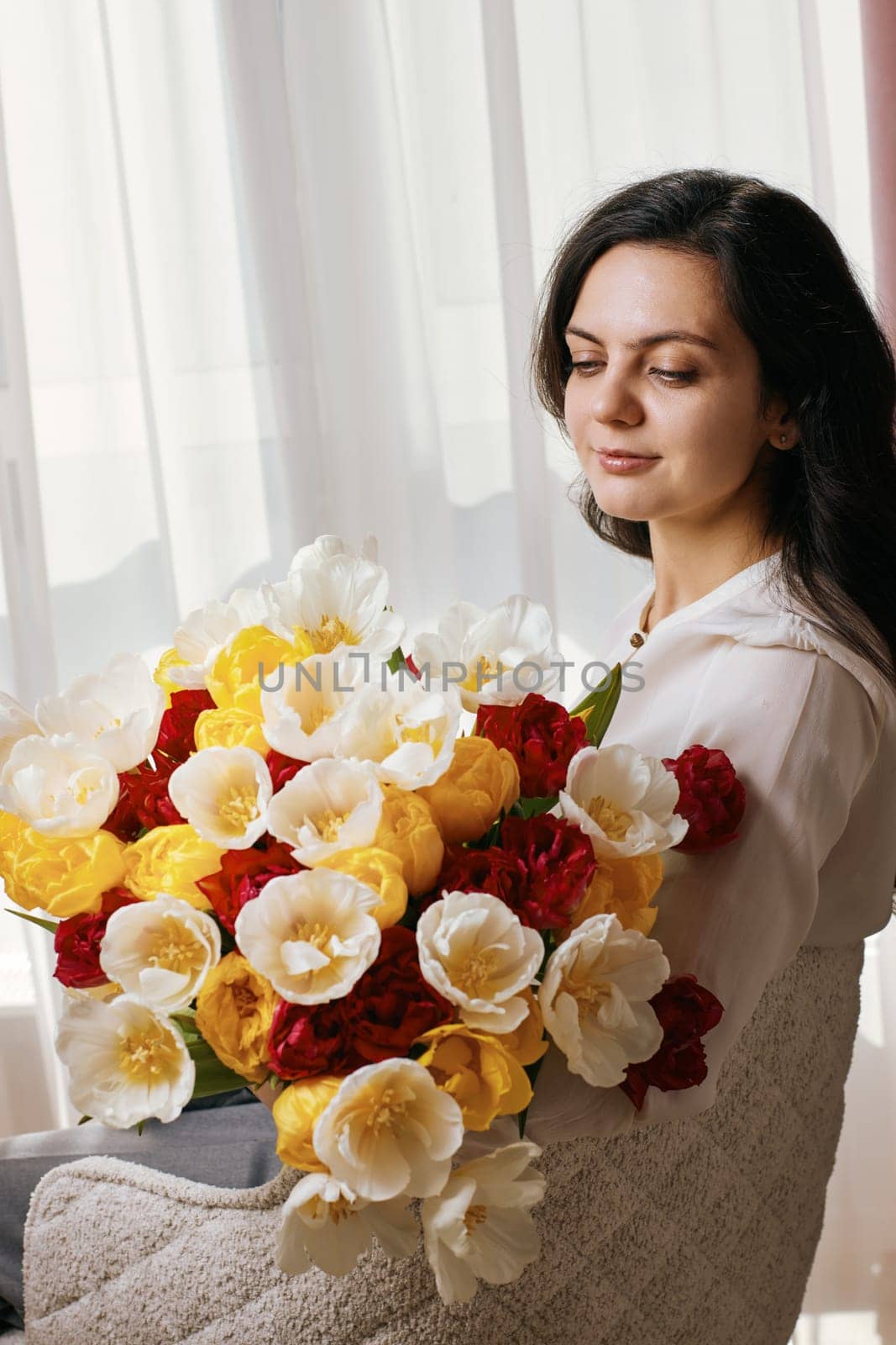 Beautiful woman with spring flowers tulips in hands sitting on chair near window. Women's Day