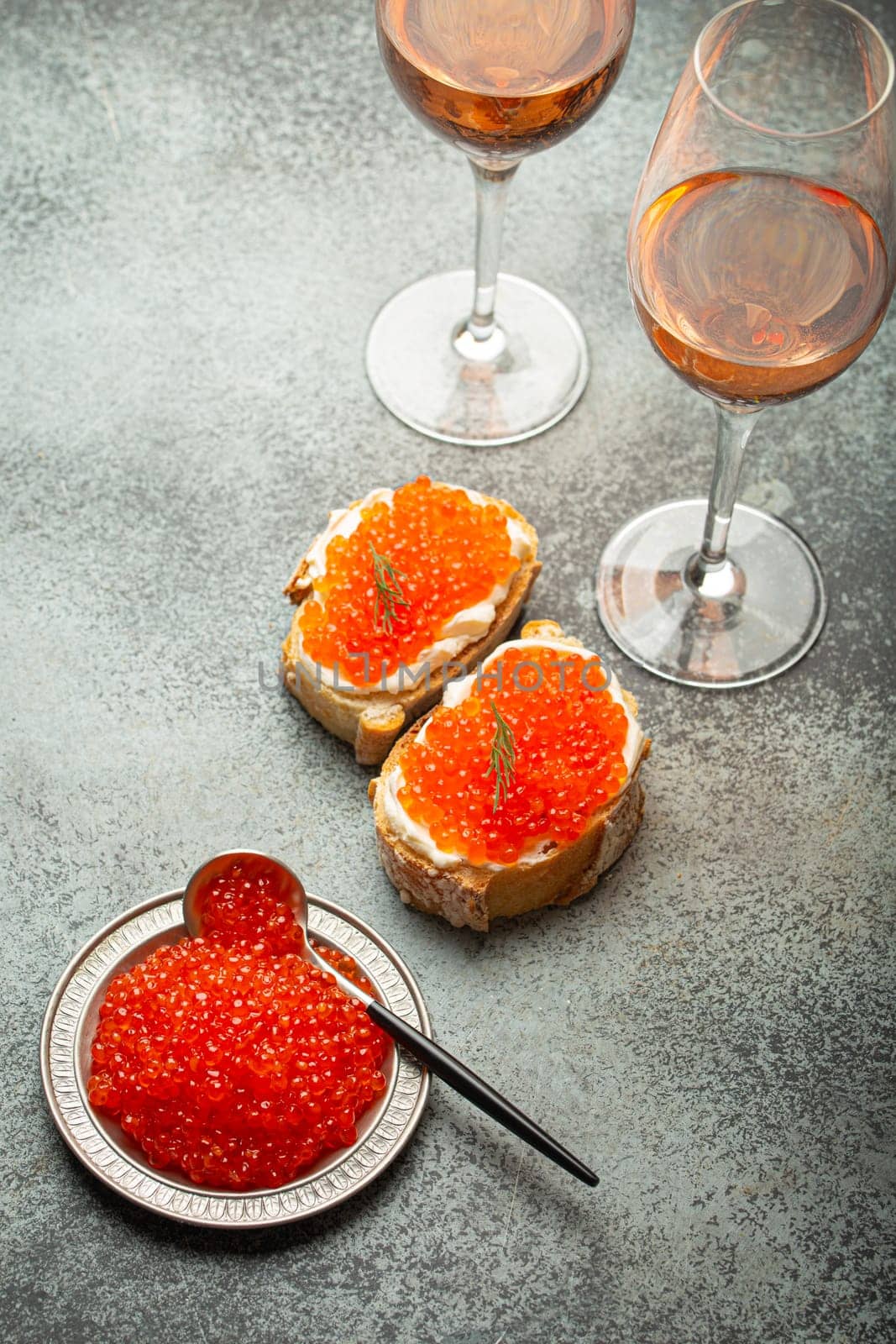 Small metal plate with red salmon caviar, two caviar toasts canape, two glasses of champagne top view on grey concrete background, festive luxury delicacy and appetizer.