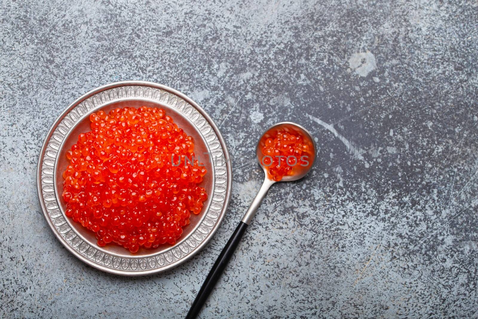 Small metal plate with red salmon caviar and a spoon top view on grey concrete background, festive luxury delicacy and appetizer by its_al_dente