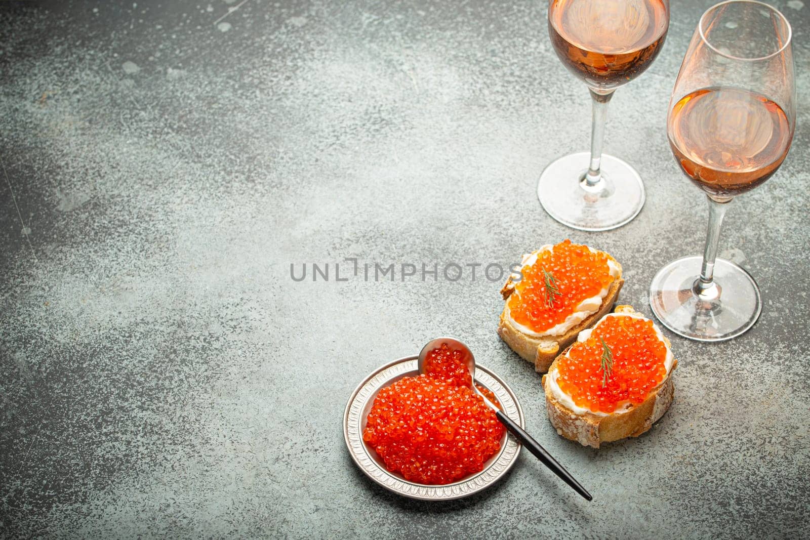 Small metal plate with red salmon caviar, two caviar toasts canape, two glasses of champagne top view on grey concrete background, festive luxury delicacy and appetizer. Space for text by its_al_dente