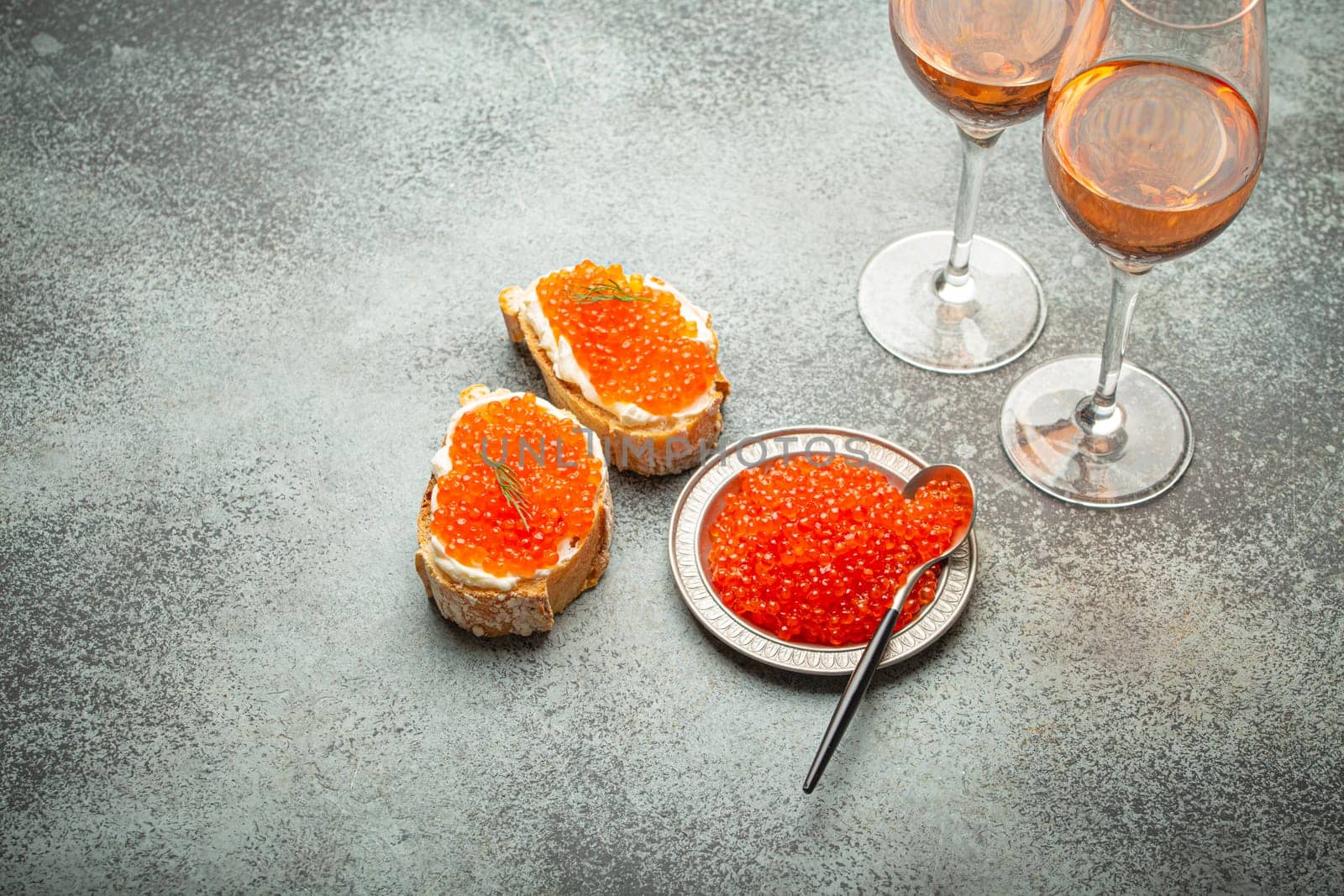 Small metal plate with red salmon caviar, two caviar toasts canape, two glasses of champagne top view on grey concrete background, festive luxury delicacy and appetizer by its_al_dente