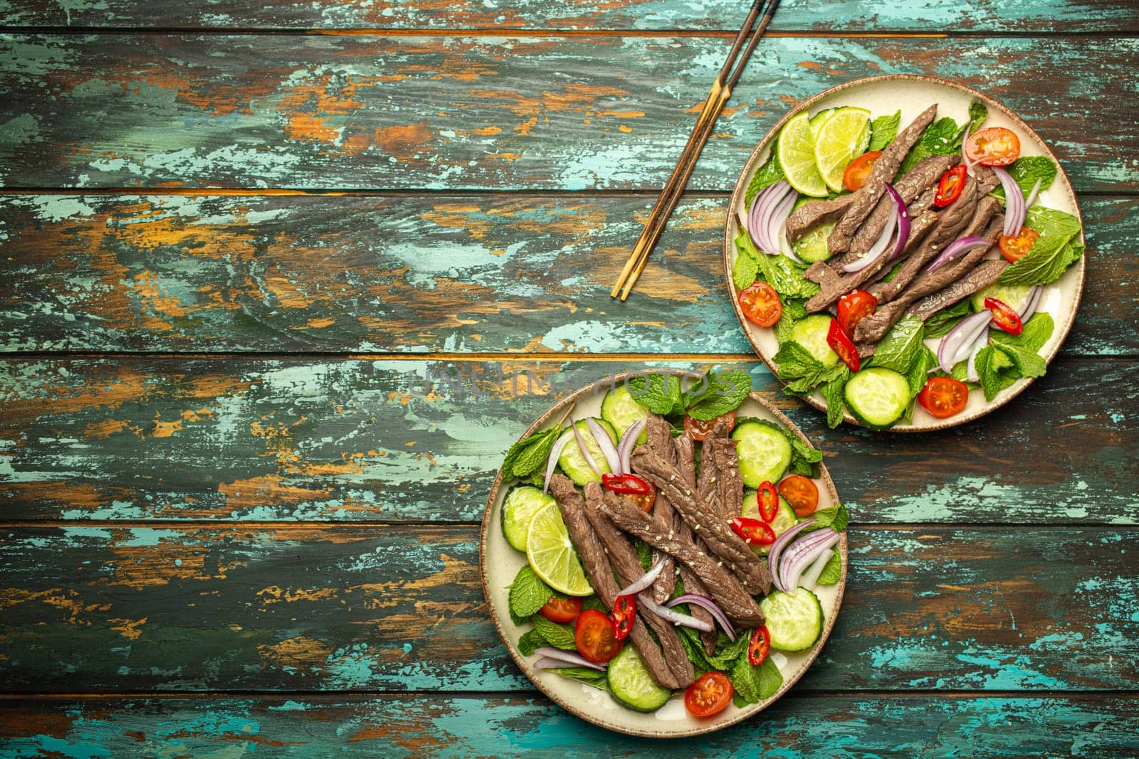 Two plates with traditional Thai beef salad with vegetables and mint top view served on rustic wooden background, healthy exotic asian meal, space for text.