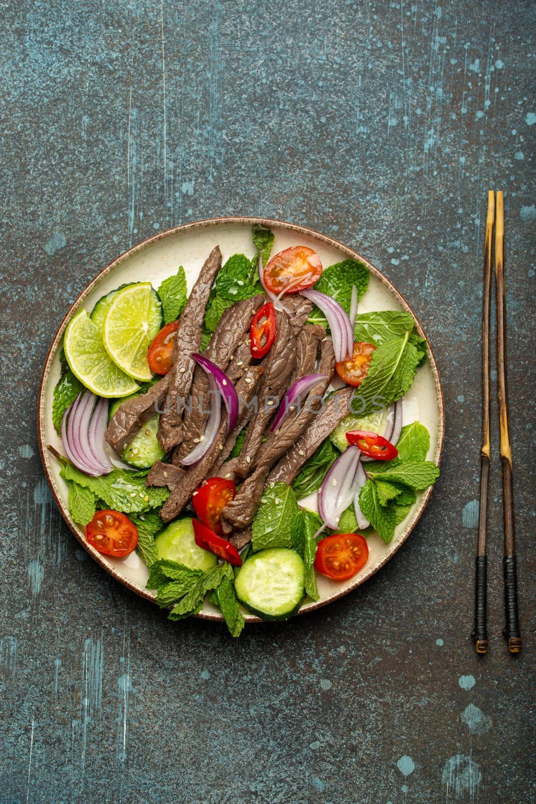 Plate with traditional Thai beef salad with vegetables and mint top view served on rustic concrete stone background, healthy exotic asian meal.