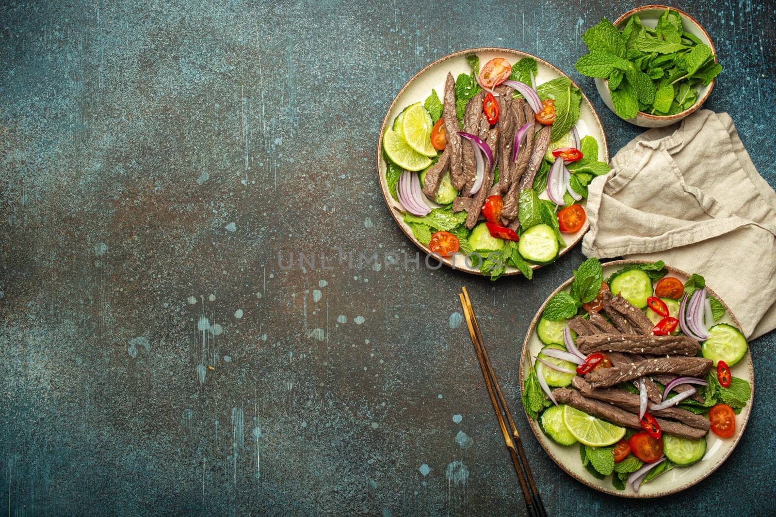 Two plates with traditional Thai beef salad with vegetables and mint top view served on rustic concrete background, healthy exotic asian meal, space for text.